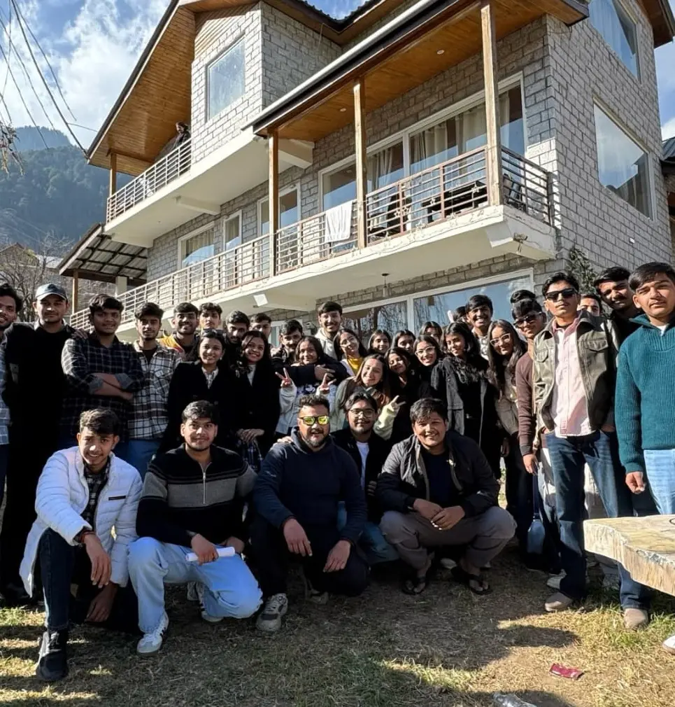 A large college group posing in front of a wooden-balconied stay in Manali on a sunny day
