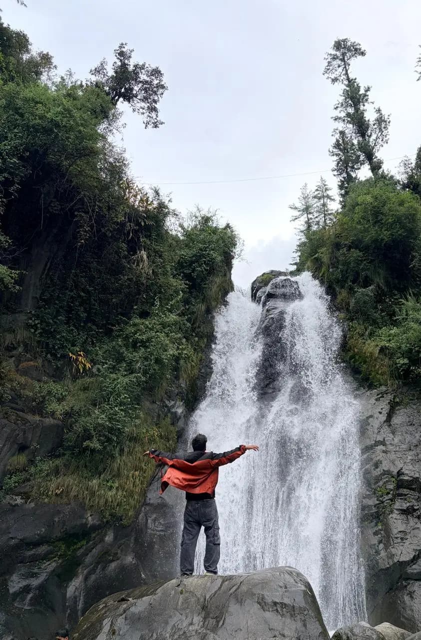 A traveller with arms outstretched on a rock in front of a tall forest waterfall
